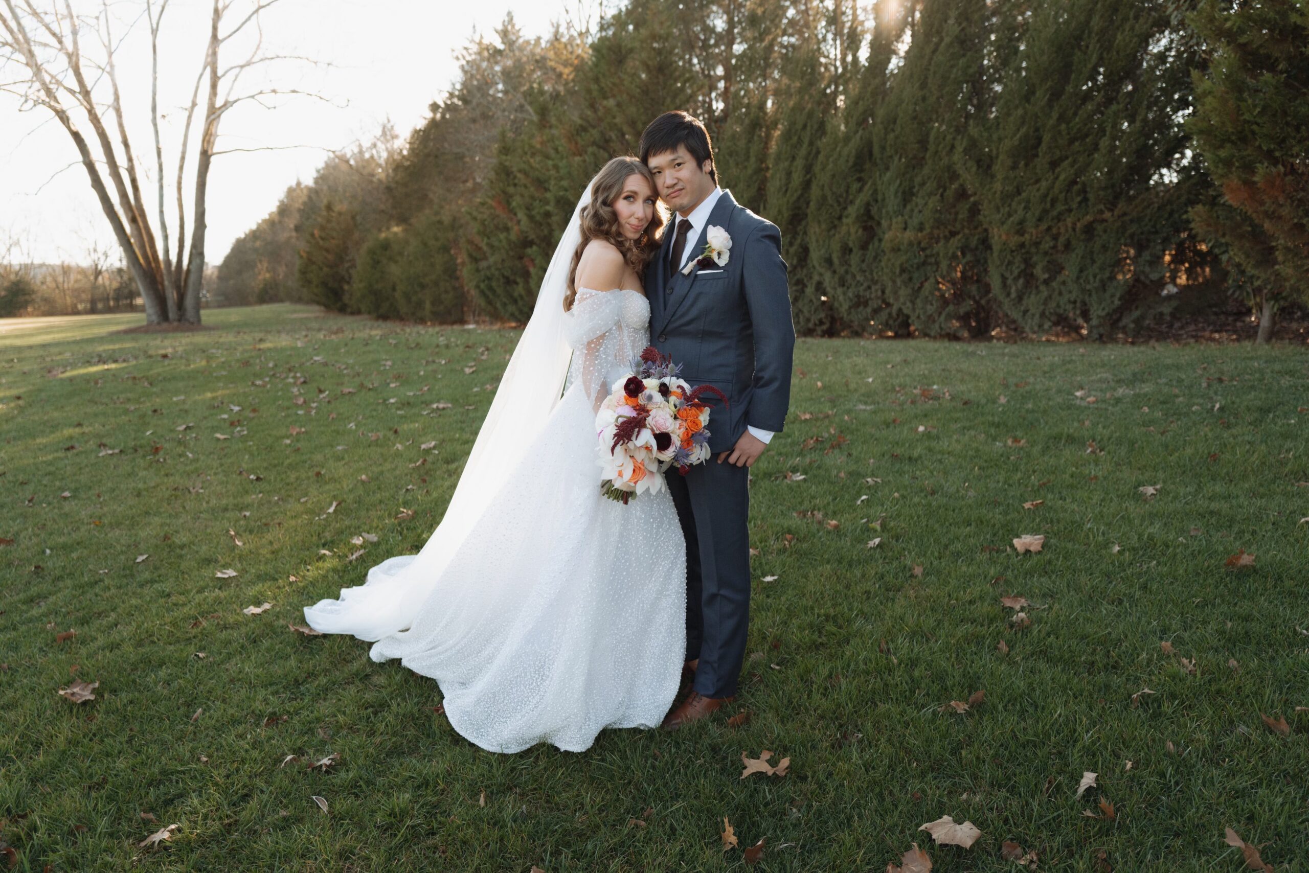 Bride and groom posing for photos in wedding attire at a partially inclusive wedding venue. Bride has white flowing dress with sparkles and is holding a bouquet. Groom is dressed in a three piece navy blue suit.