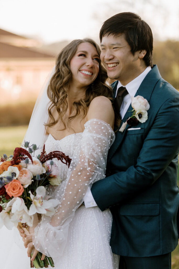 Bride smiling and looking at her groom while he cradles her from behind for a photo.