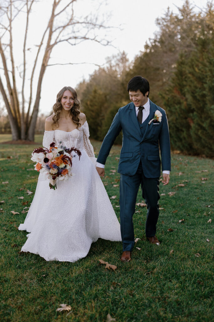 Bride and groom walking hand in hand with the bride holding a large and colorful bouquet. The bouquet has peach, burgundy, and pink flowers. 