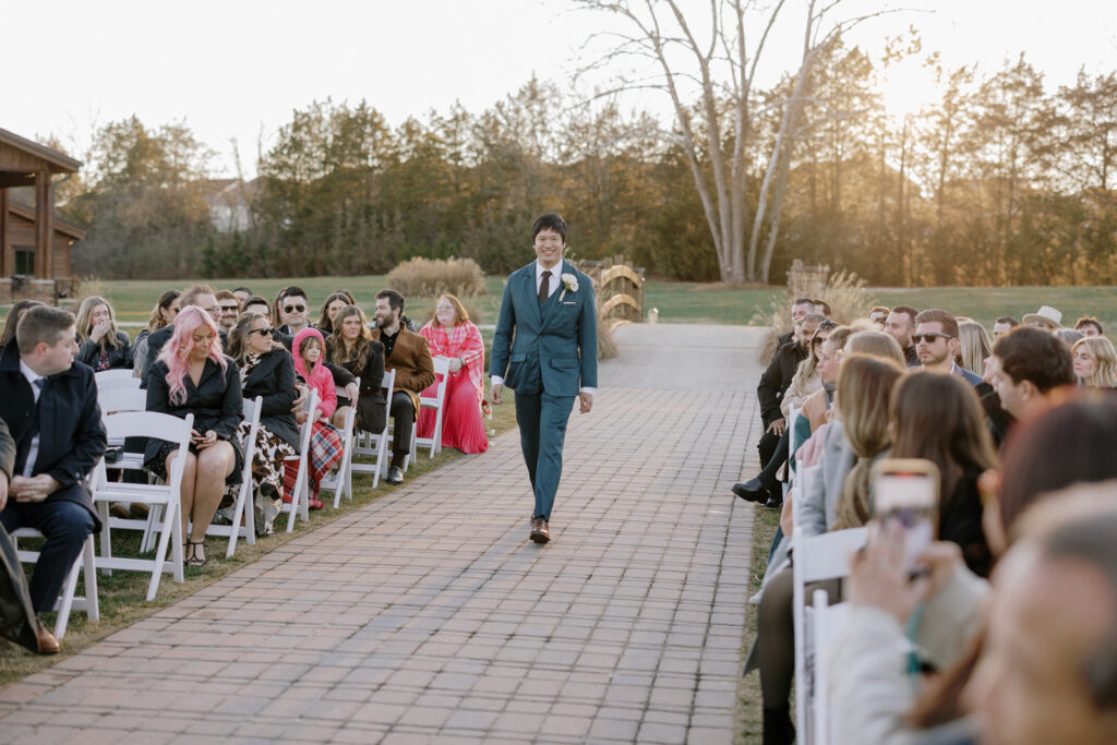 Groom walking down the aisle for his ceremony. He is wearing a navy blue three piece suit and smiling.