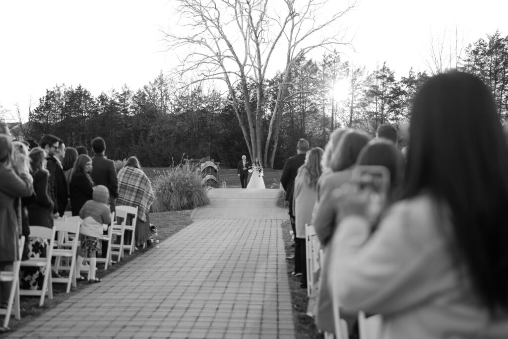 A black and white photo of the bride walking with her father down the aisle to the wedding ceremony. 