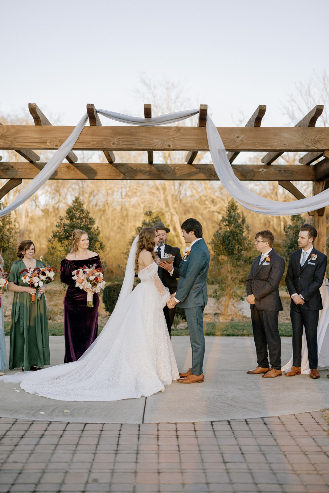 Bride and groom stand facing each other at the ceremony under a wooden pergola.