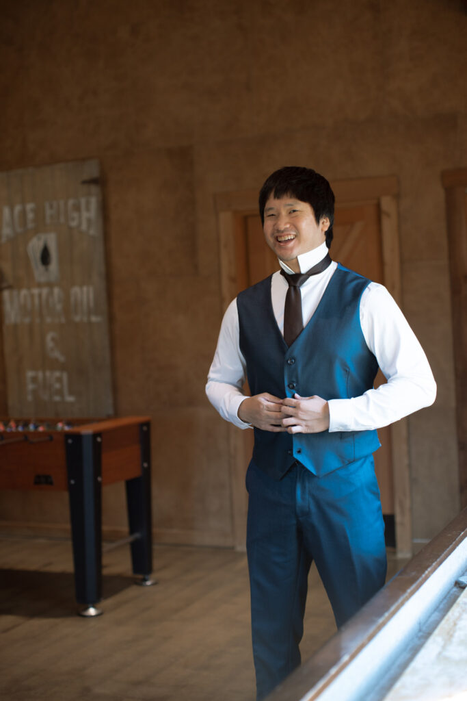 Groom in suite at Sycamore Farms buttoning vest of his custom navy blue suit and smiling. 