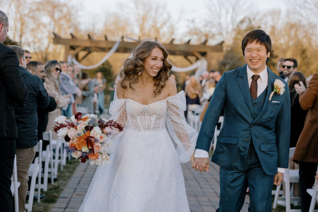 The bride walking out of the ceremony with her groom after being married. She has a big smile on her face and his holding his hand. 