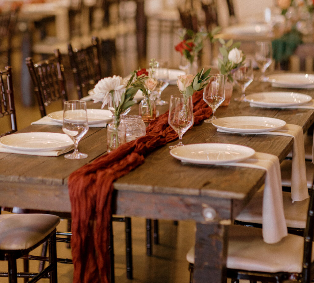 Table at a wedding with rust red runner, white plates, white linen napkins, and water glasses.