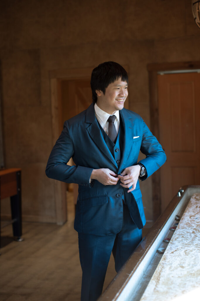 The groom buttoning his navy blue suit jacket looking out the window and smiling. 