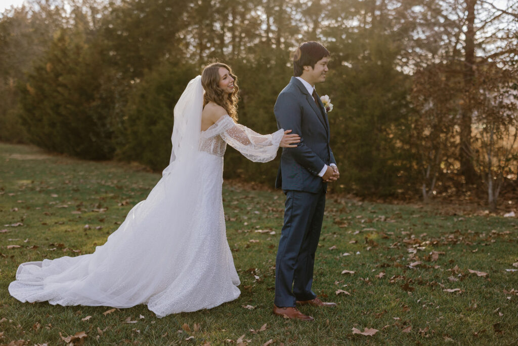 Bride approaching groom who has his back turned waiting to see his bride for the first time. They are outside in a green grass lawn with trees behind them.