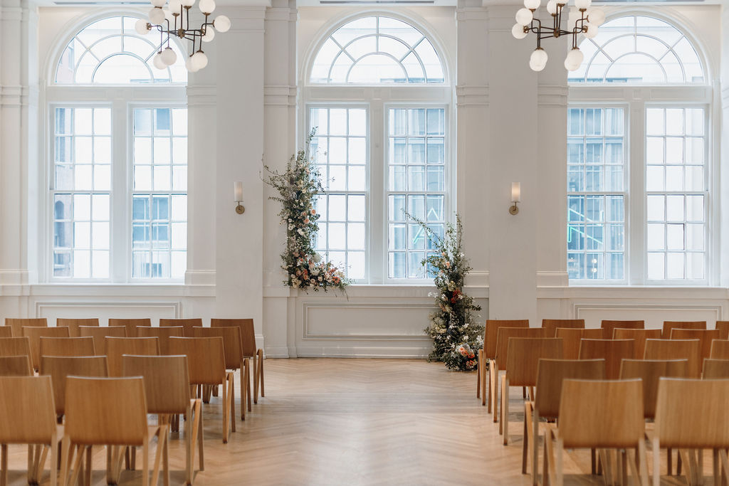 Ceremony site with 3 large windows. Center window has floral arch arrangement. Wooden chairs are in rows facing floral arrangement and windows with a center aisle.