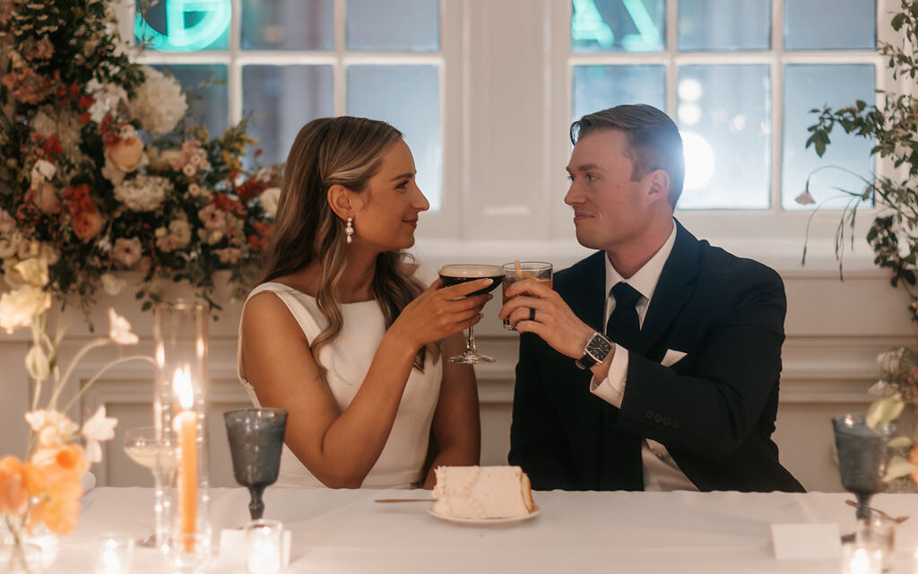 Bride and groom face each other while seated at their wedding reception. They are smiling at each other and clinking glasses in celebration.