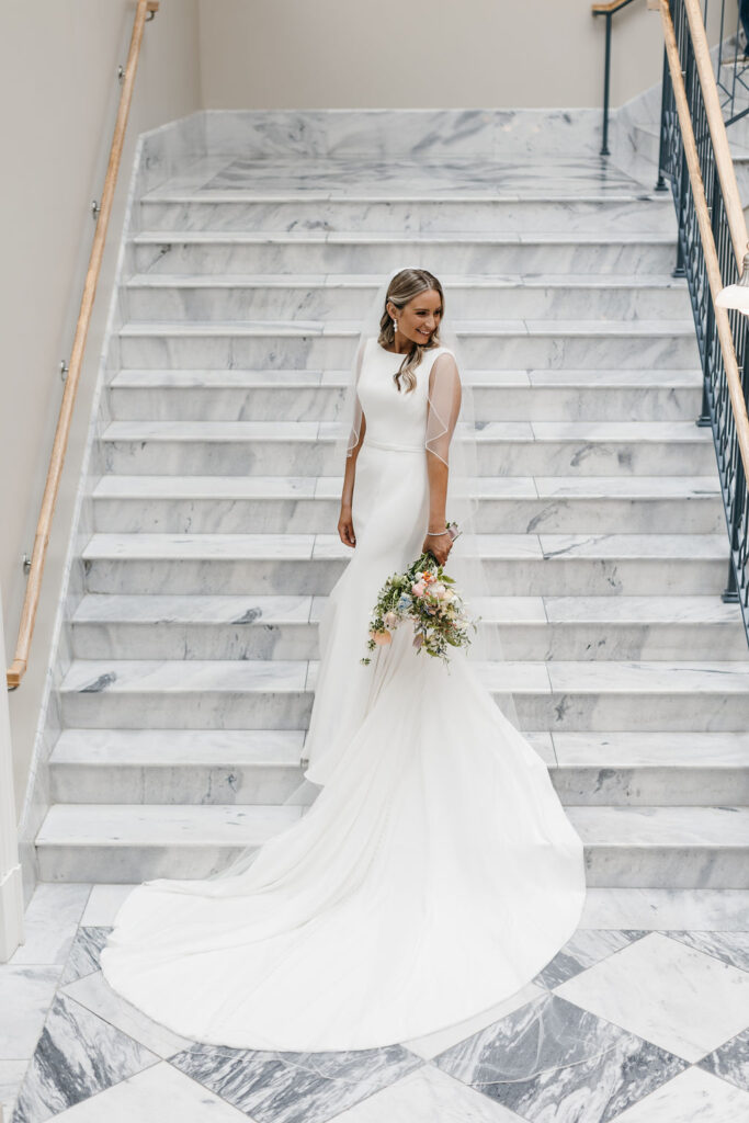 A bride in a long white dress with veil standing on a marble staircase looks to her left and smiles