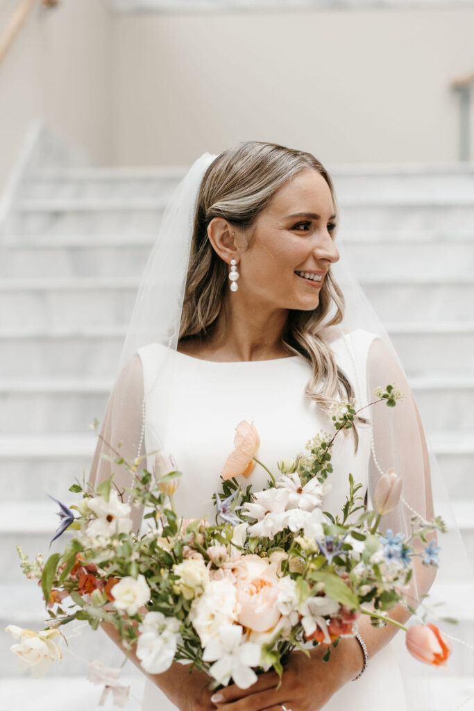 A bride in a long white dress with veil standing on a marble staircase looks to her left and smiles