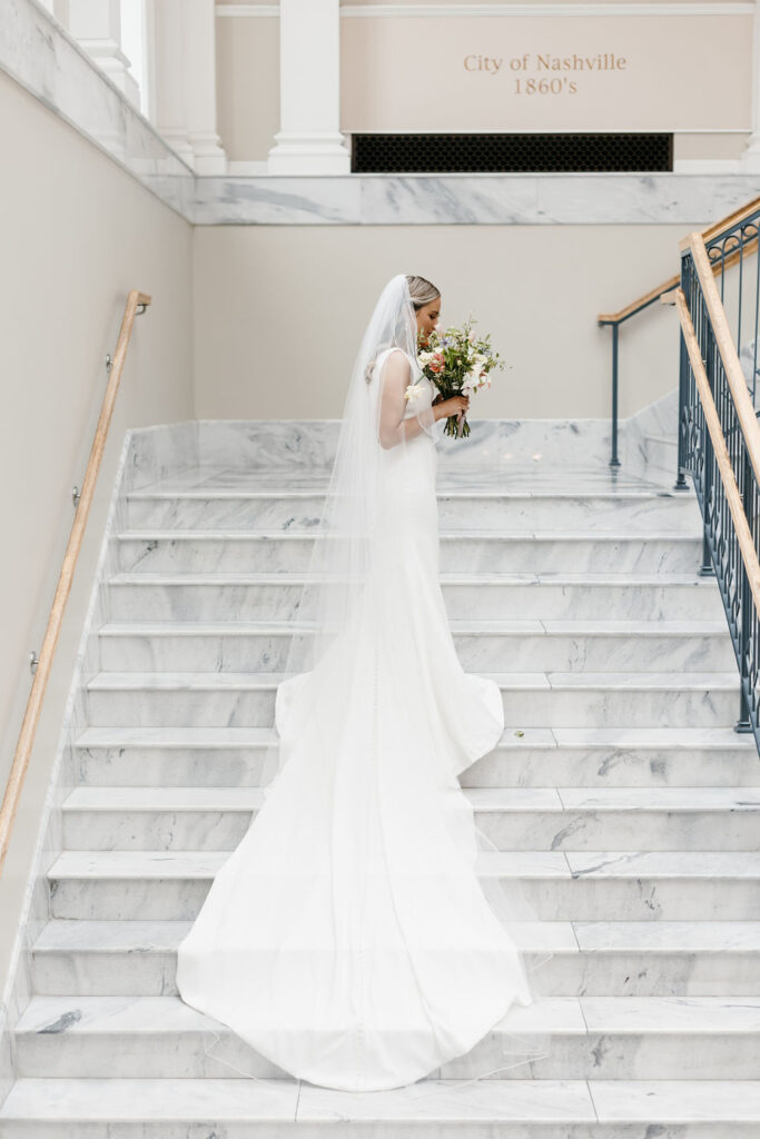 A bride in a long white dress with veil standing on a marble staircase smells her bouquet of flowers