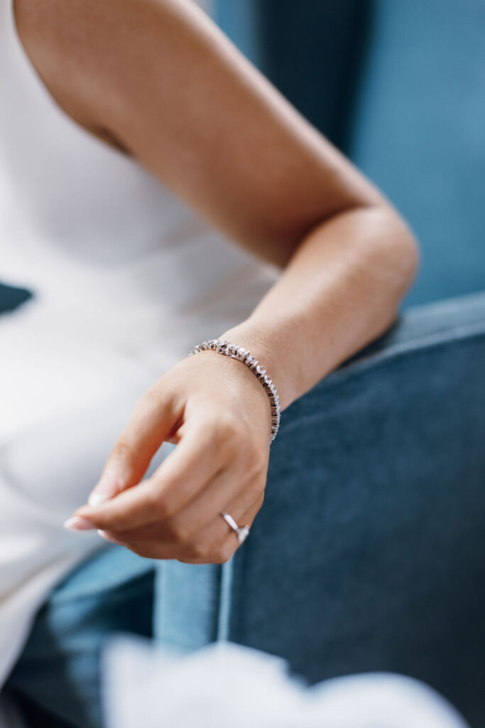 A detail photo of a diamond bracelet and diamond ring as the bride sits in a teal armchair.