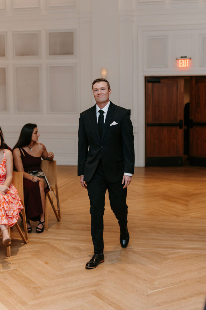 A groom in a black suit walks down the aisle to his wedding ceremony