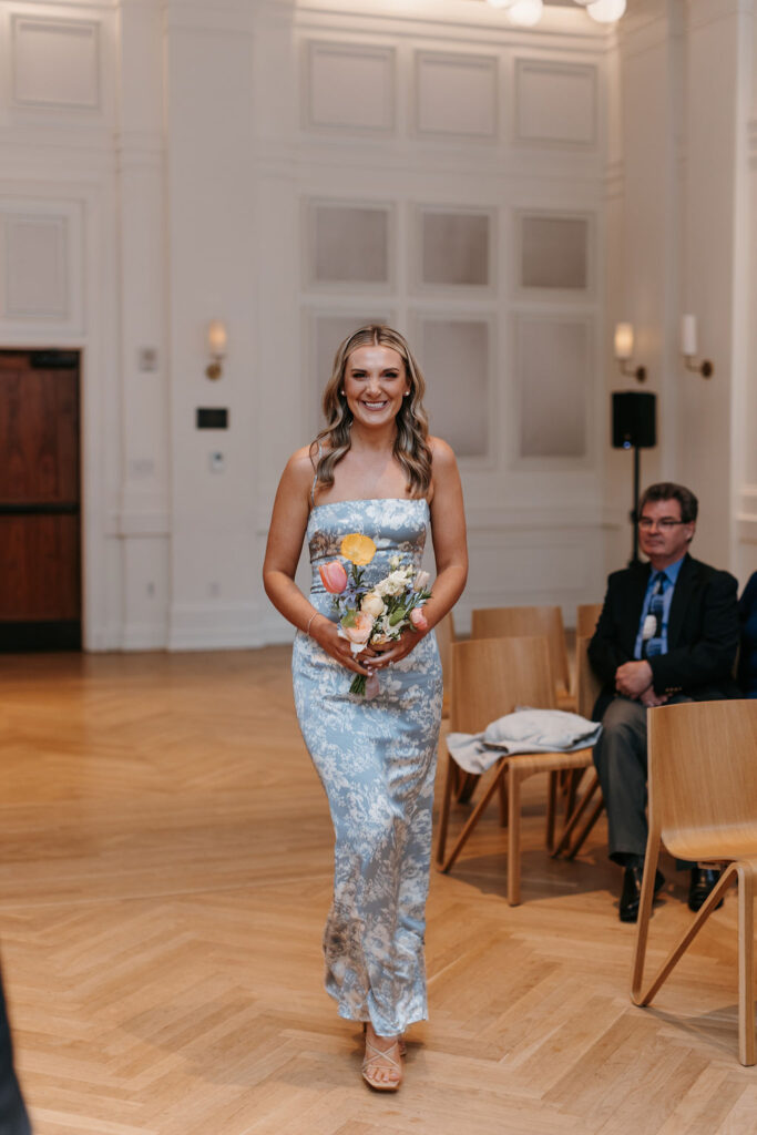 The maid of honor walks down the aisle in a blue bridesmaid dress carrying a colorful floral bouquet and smiles