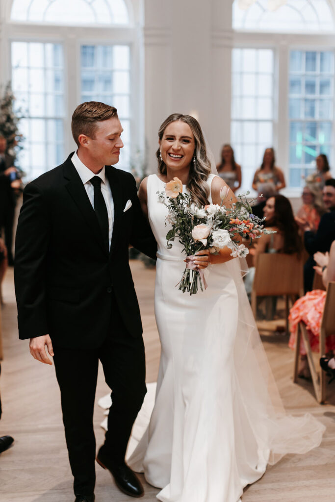 A bride and groom laugh playfully as she carries her colorful bridal bouquet while walking down the aisle