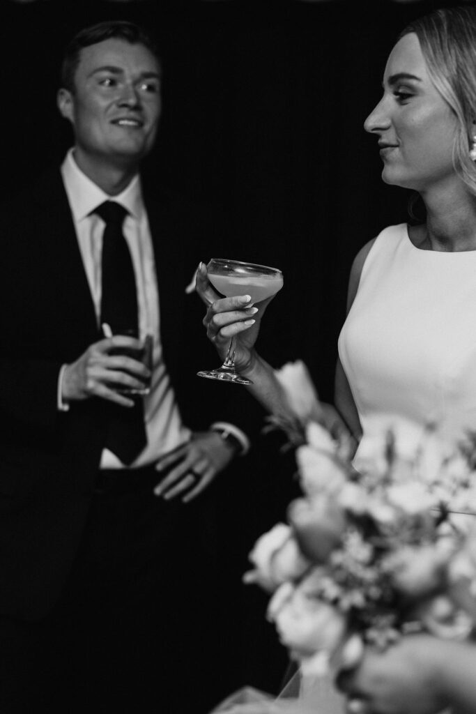 A groom looks happily at his bride as they drink cocktails