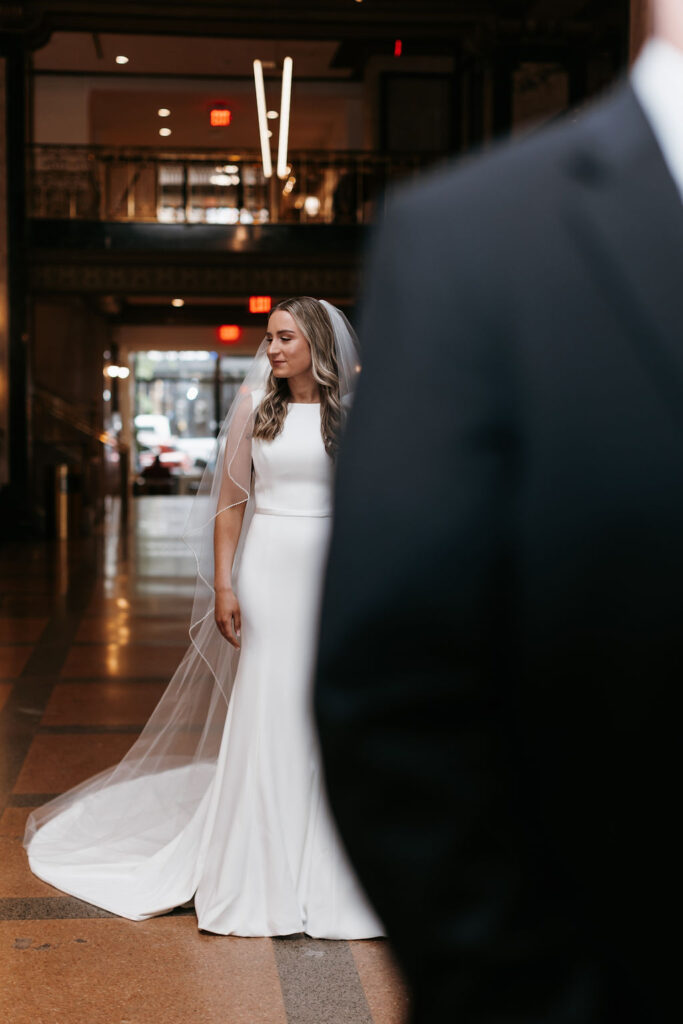 A bride in her white dress and veil looking to the side as she stands in the background of the camera