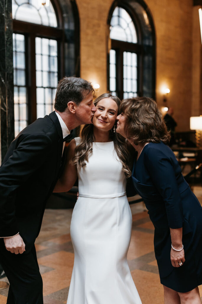 The mother and father of the bride both kiss their daughter on the cheek