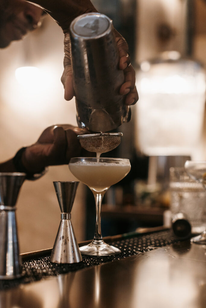 A bartender pours a cocktail from the shaker into a glass coupe