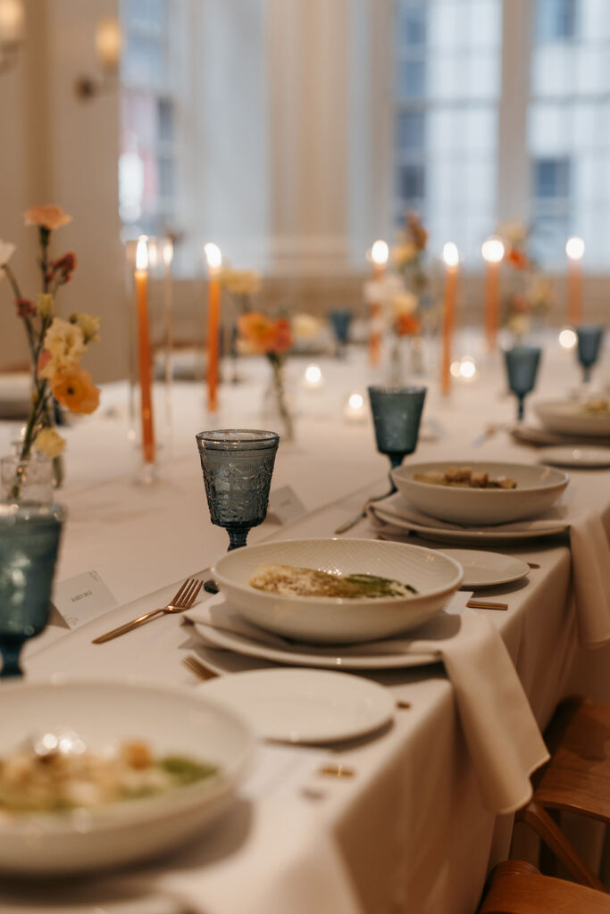 Wedding reception tables with orange taper candles and orange flowers set with blue water glasses and a caesar salad