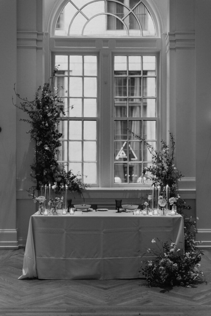 A table set for the bride and groom with glowing candles and a floral installations behind in the window