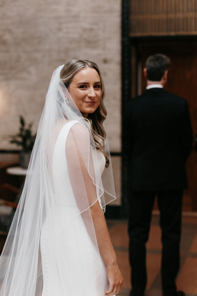 A bride turns to look behind her with a small smile on her face before revealing her outfit to her father