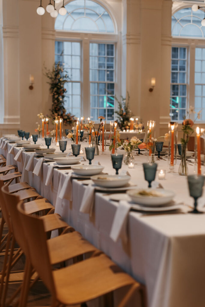 Wedding reception tables with orange taper candles and orange flowers set with blue water glasses and a caesar salad