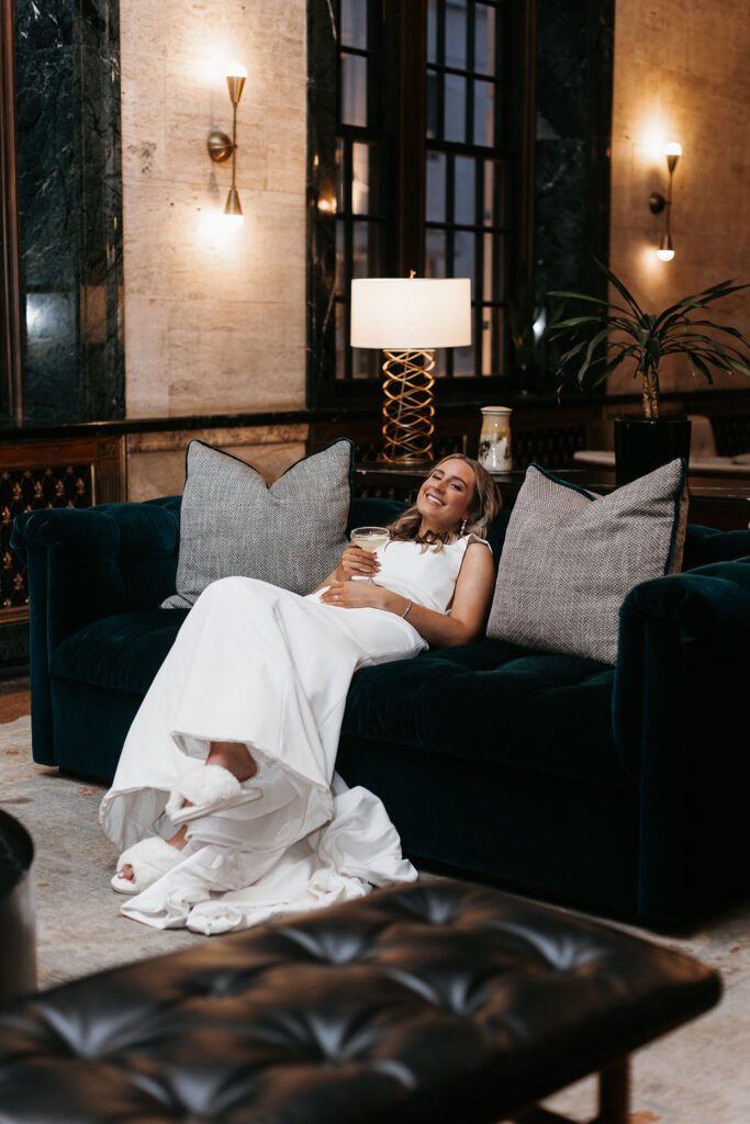 A bride lounging on a couch with a cocktail coupe in her hand as she wears her wedding dress and white fluffy slippers