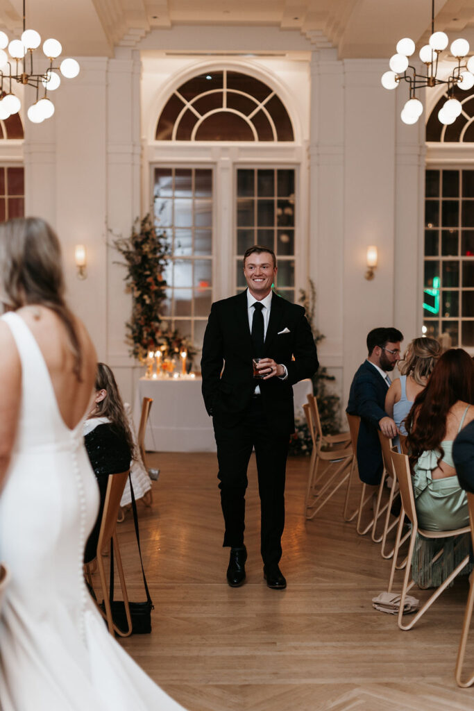 A groom in a black suit at the wedding reception admiring his bride who is talking with guests