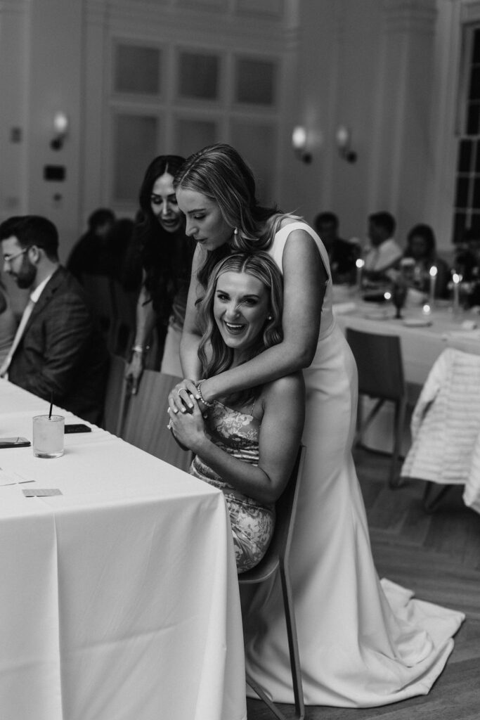 A bride hugging her friend from behind as her friend sits in the chair and smiles at the camera