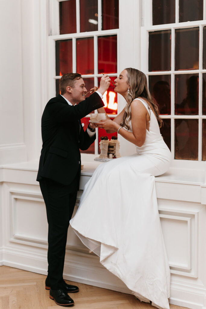 A bride sits on a windowsill as the groom feeds her cake