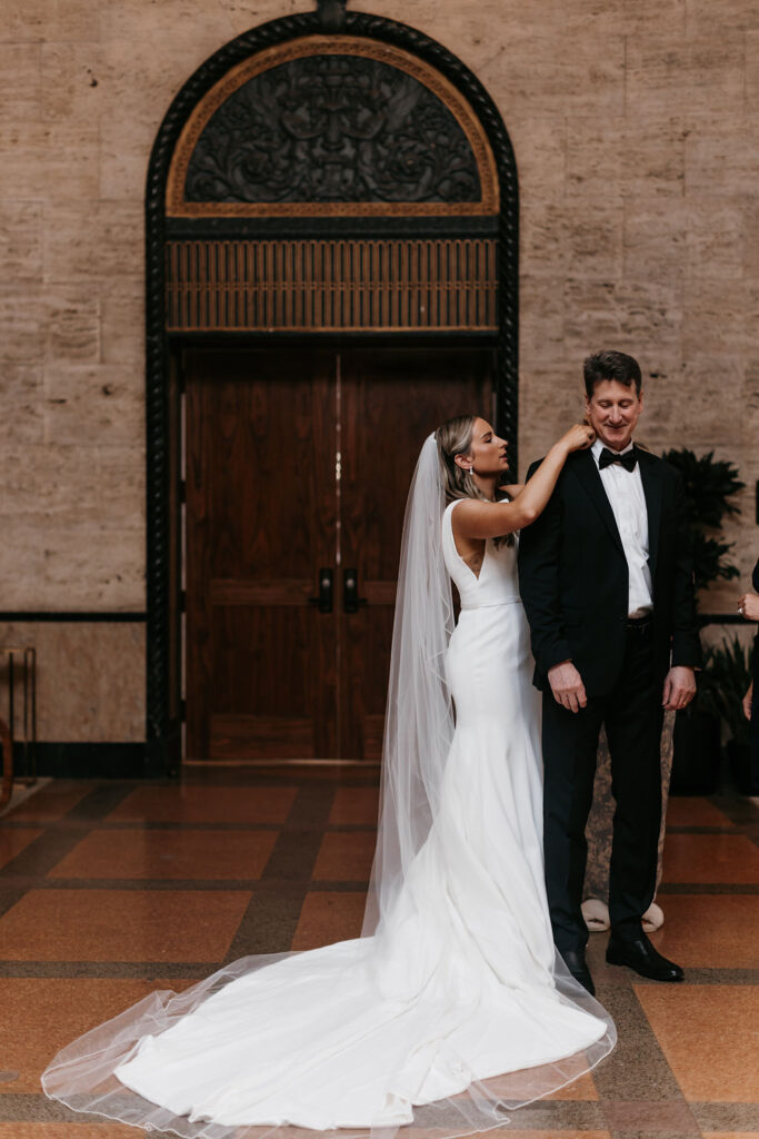 The bride stands next to her father and adjusts his black bowtie to match his two piece black suit