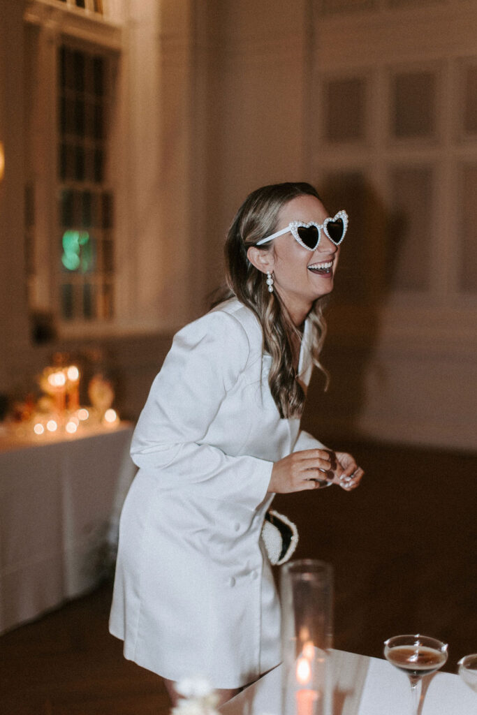 A bride wearing white heart shaped sunglasses indoors as she laughs off to the side