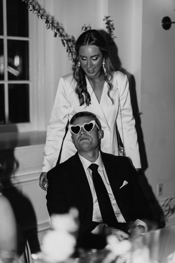 A groom sitting and wearing white heart shaped sunglasses inside during a wedding reception