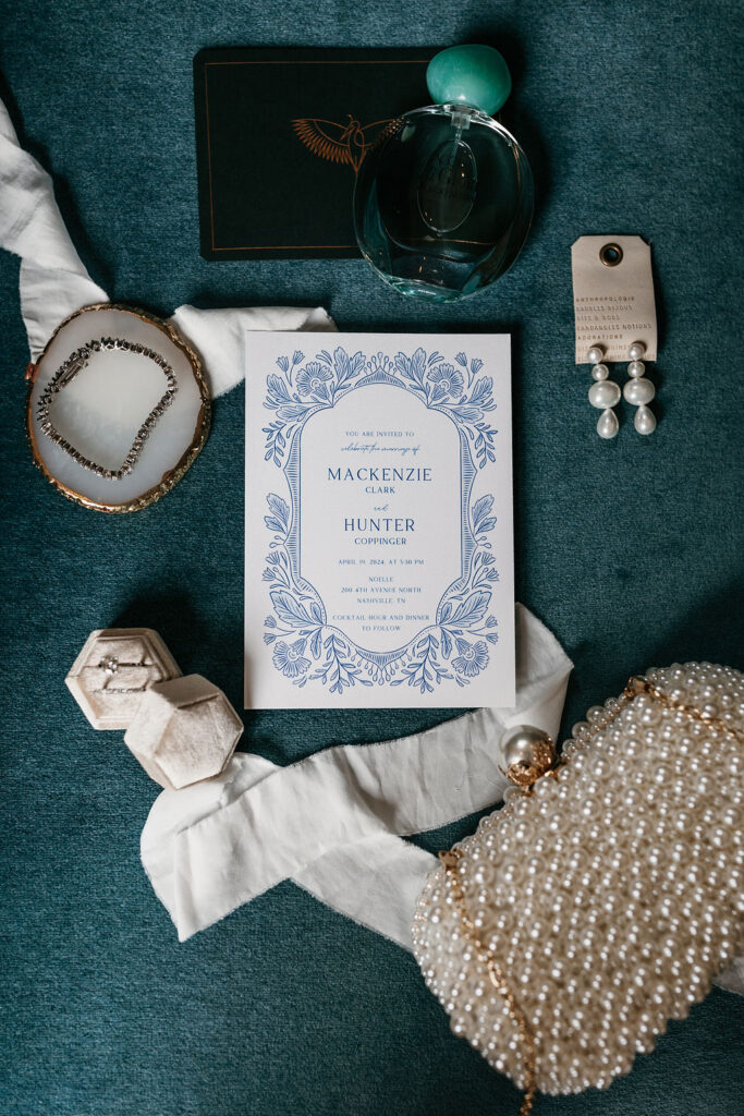 Blue wedding invitations surrounded by bridal accessories including diamond bracelet, wedding ring, pearl earrings, and pearl covered clutch