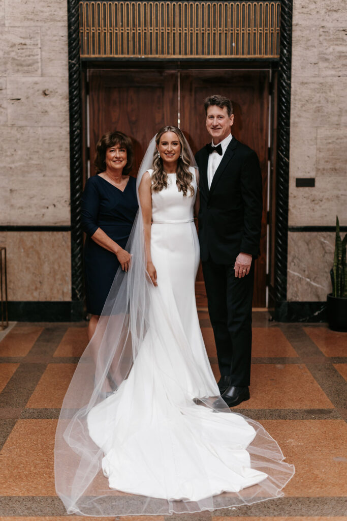 A mother, father, and daughter pose for a photo together on the daughter's wedding day