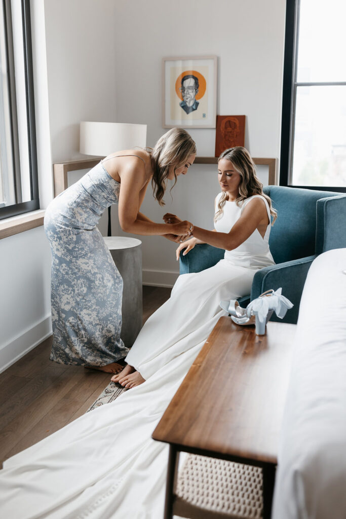 A bride sitting in a teal armchair as her maid of honor assists her in accessorizing with a diamond bracelet