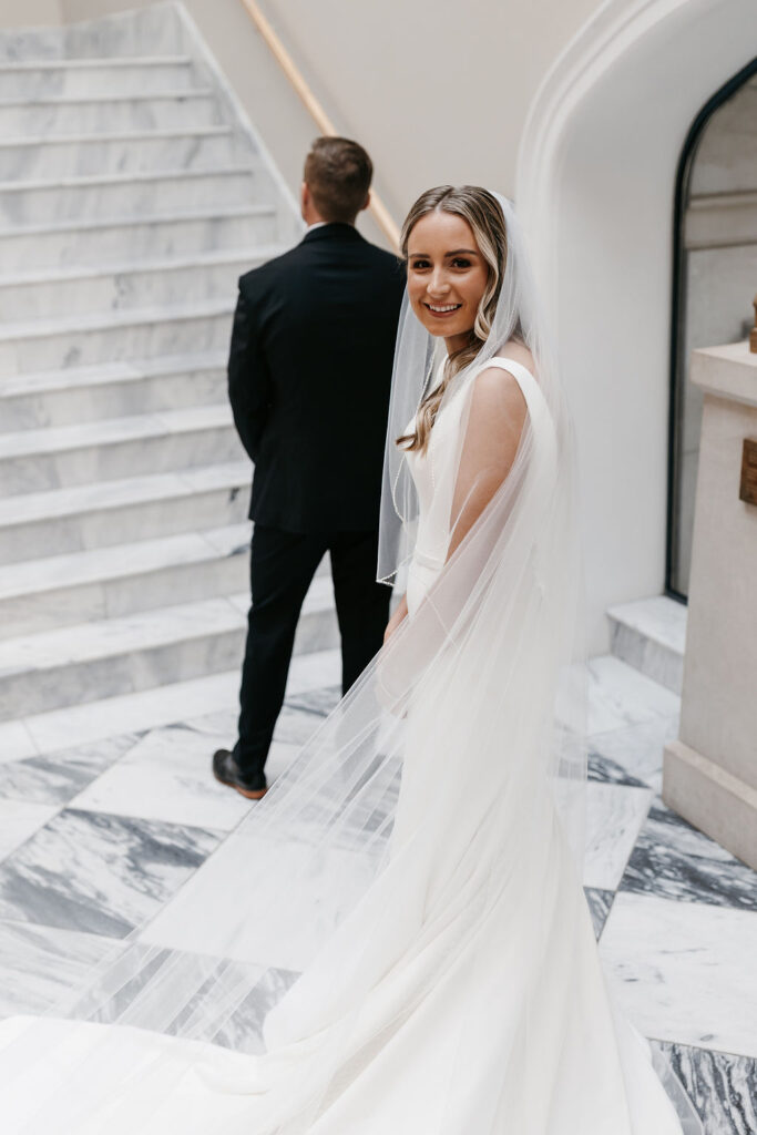 A groom stands before a marble staircase as the bride turns to the camera with a smile