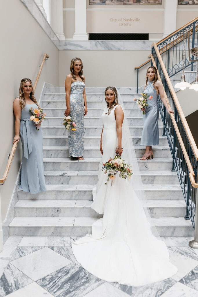 Three bridesmaids in blue dresses carry bouquets as they stand on alternating steps on a marble staircase with the bride in a long white dress