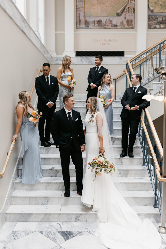 A wedding party in blue dresses and black suits stand at different steps on a marble staircase with the bride and groom in front