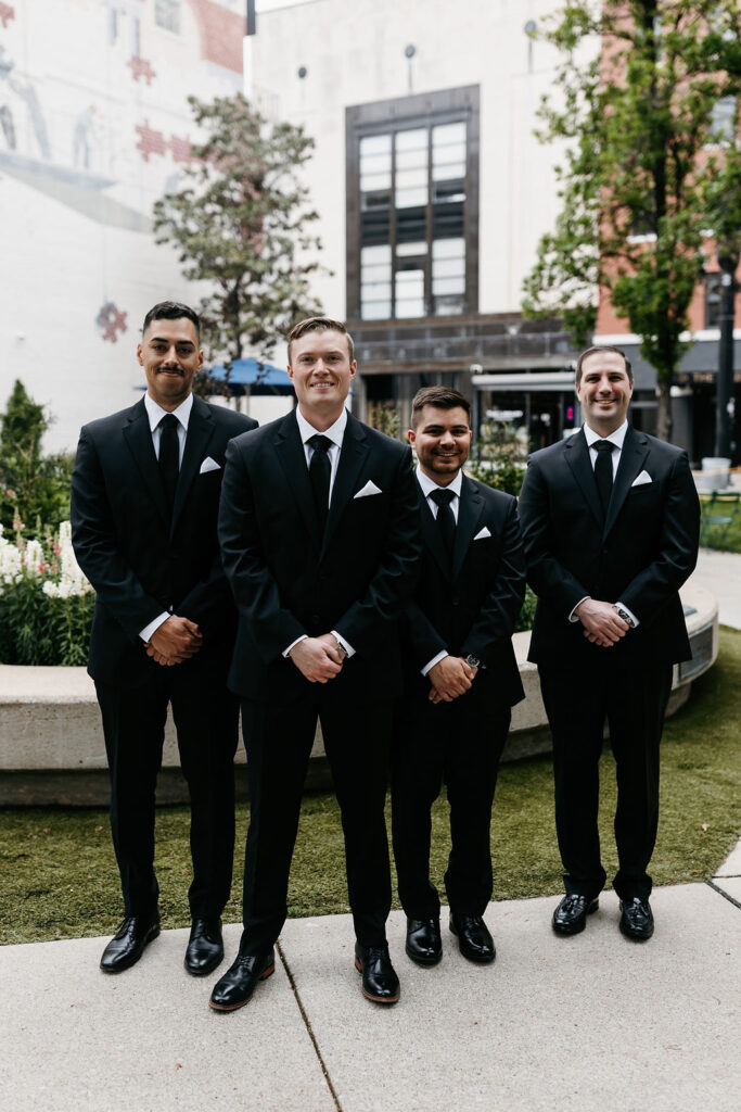 Four groomsmen in black suits stand side by side in front of a fountain