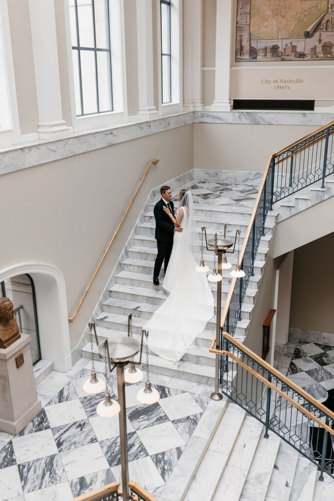 The bride and groom stand on a marble staircase in the light filled Nashville Public Library main branch