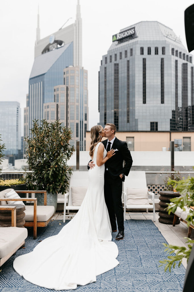 A bride and groom hug each other and kiss on a hotel rooftop with the Nashville downtown skyline behind them