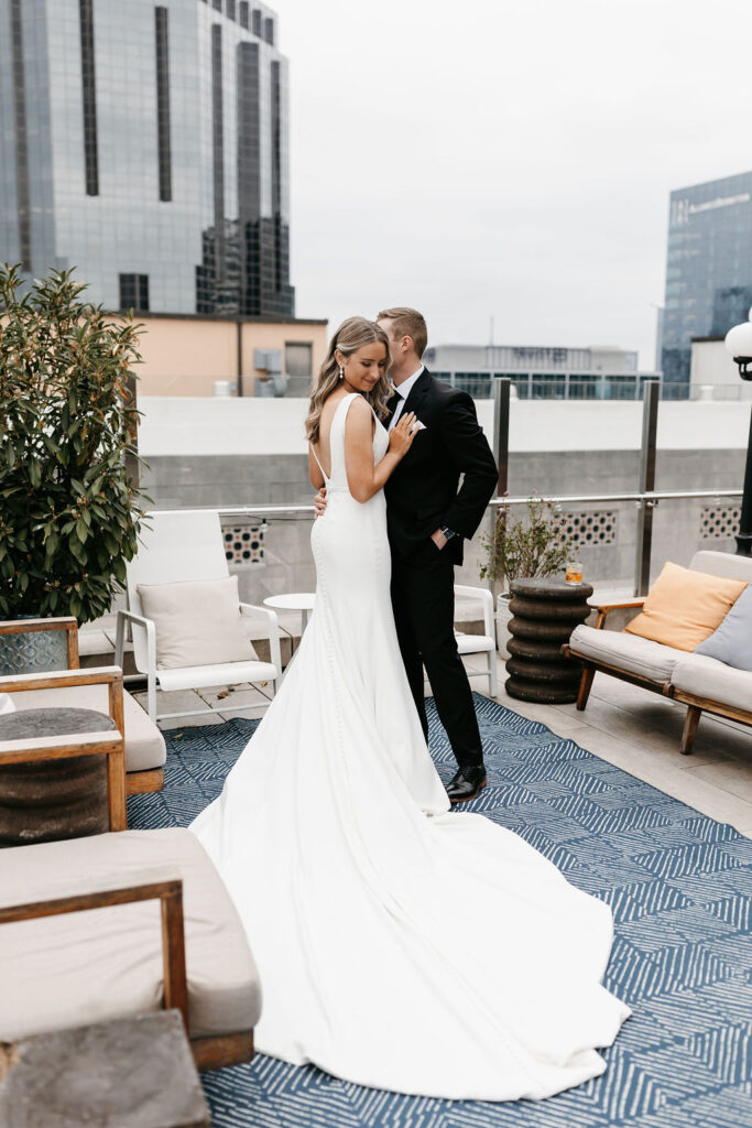 A bride and groom hug each other and kiss on a hotel rooftop with the Nashville downtown skyline behind them