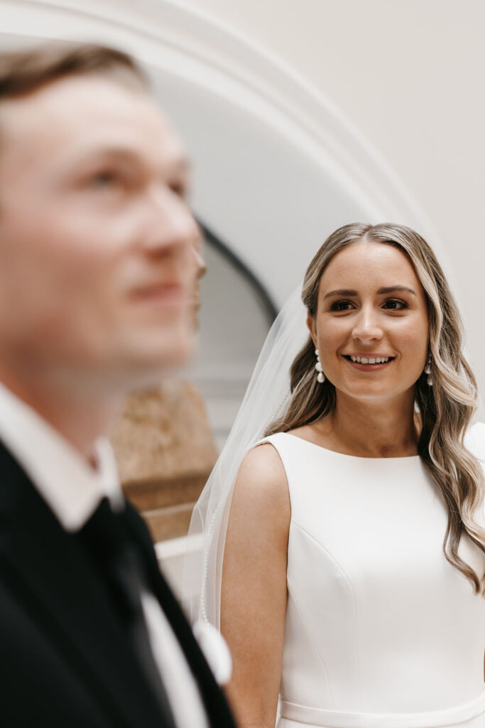 The groom is unfocused in the foreground as the bride who is in focus smiles at her groom