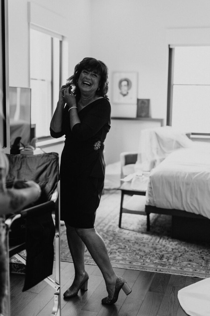 Mother of the bride posing in a hotel room with a big smile on her face while she puts on earrings