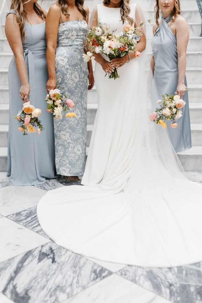 Three bridesmaids in blue dresses carry bouquets as they stand side bt side on a marble staircase with the bride in a long white dress