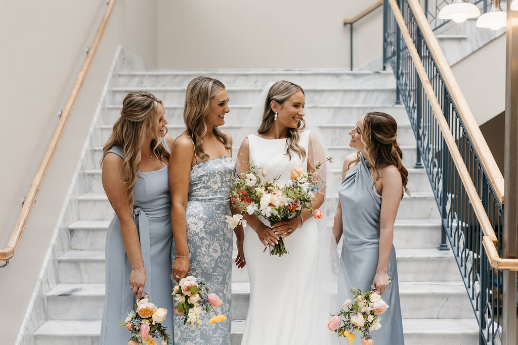 A bride at her wedding smiles at a bridesmaid in a blue dress carrying a bouquet in front of marble steps