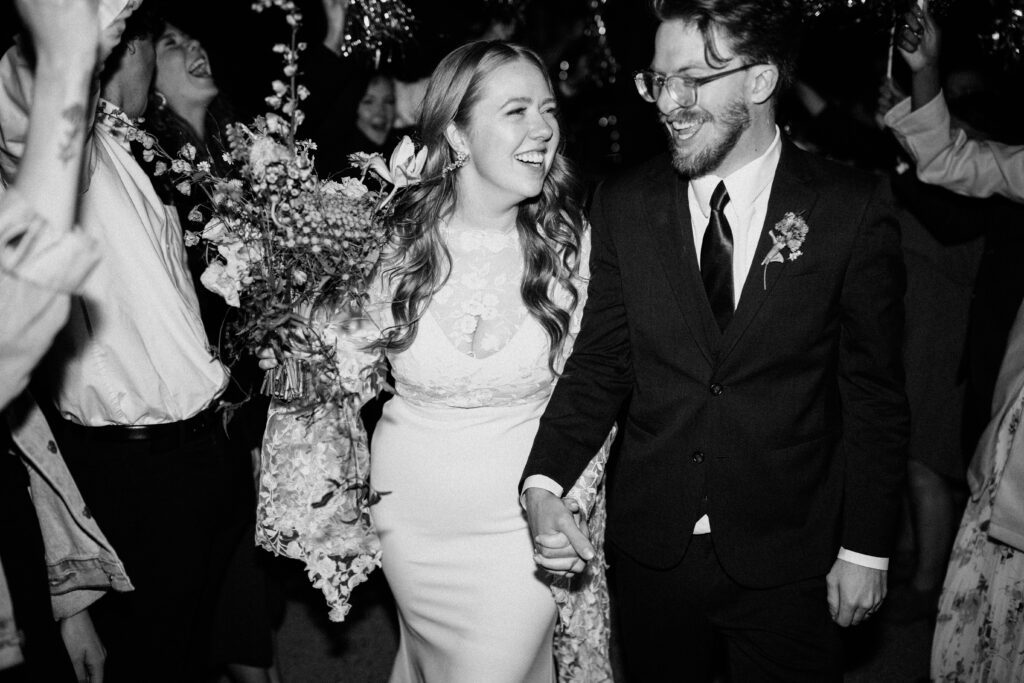 bride and groom smile and hold hands with their wedding guests cheering around them